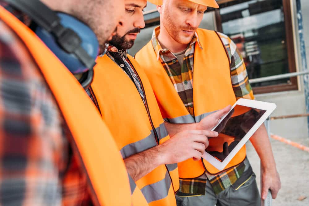 Construction team in high-visibility vests and hard hats collaborating while inspecting a digital plan on a tablet, showcasing Smartway Builders' commitment to modern methods and teamwork.